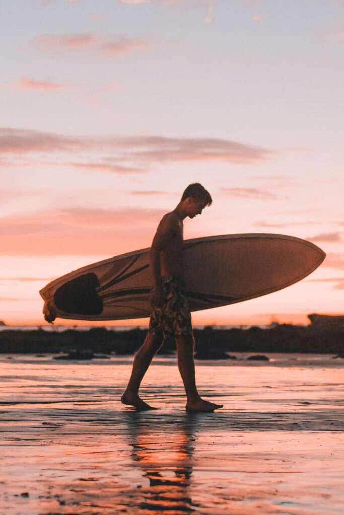 Man with surfboard walking along the beach during sunset.